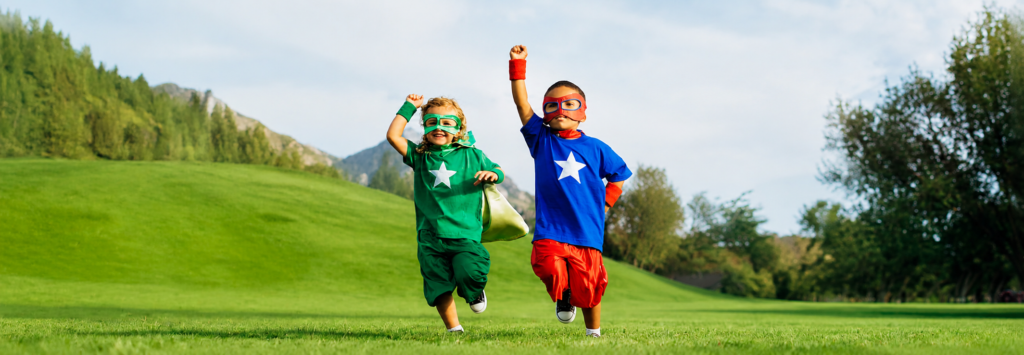 Two children in superhero costumes run across a sunny grassy field, fists raised in triumph.