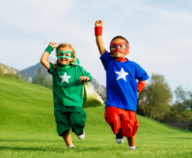 Two children in superhero costumes run across a sunny grassy field, fists raised in triumph.